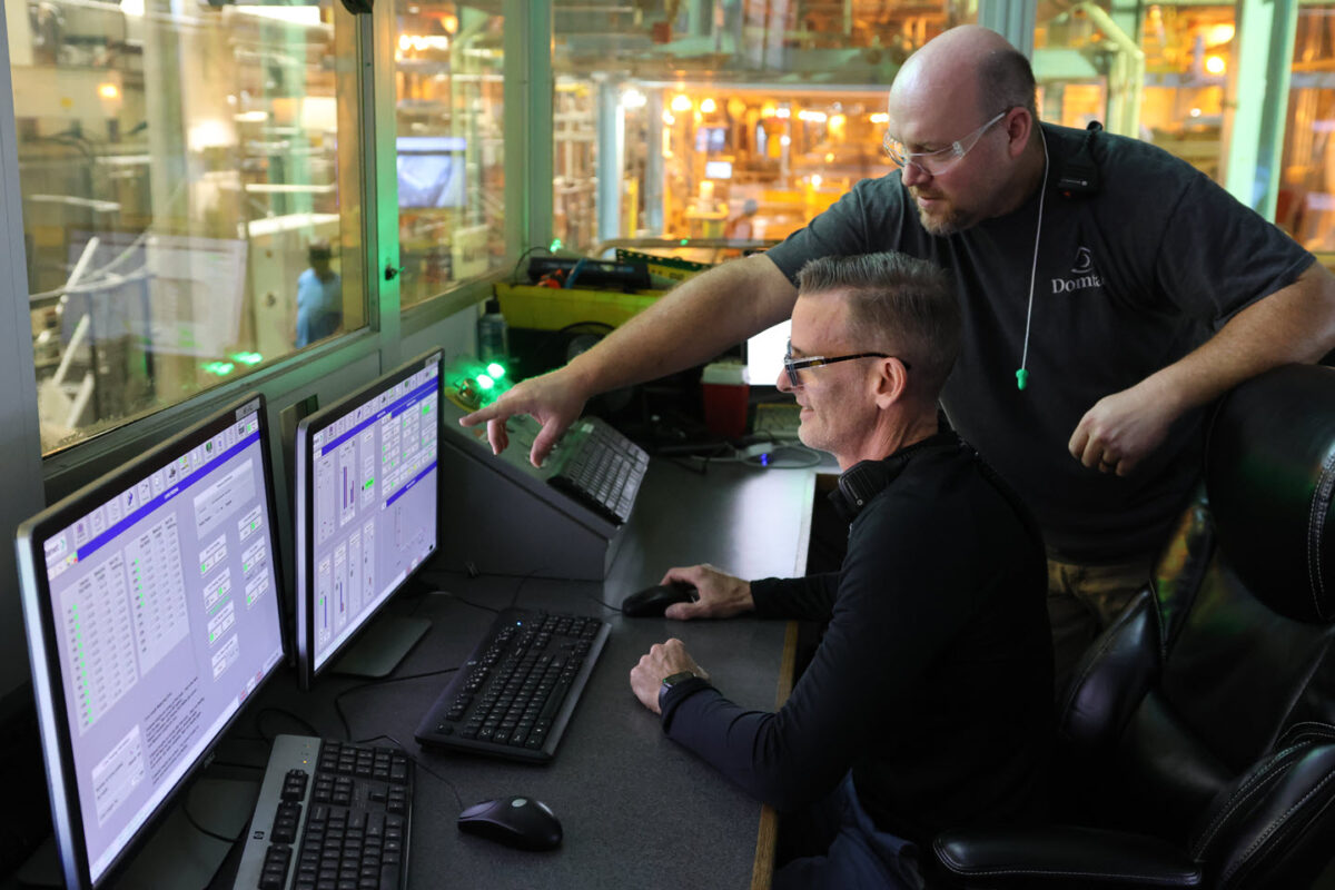 Two male workers in a control room at a pulp mill are looking at a computer screen. The man in the back is standing and pointing something out on the screen to the other man, who is seated. Institutional knowledge is being shared to help each other do their jobs.